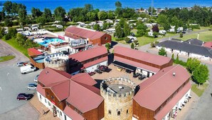 En slottsliknande byggnad med ett centralt torn, omgiven av mindre byggnader med röda tak, och i bakgrunden syns ett kustlandskap med klar himmel.