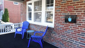 A covered outdoor seating area with two blue chairs and a small table, a brick wall, a window with white frames, and a mailbox with a blue and white logo. - Serene modern 3-bedroom house in Shillington (Shillington)