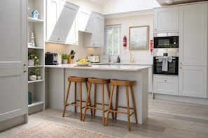 Une cuisine moderne avec des meubles blancs, un îlot central doté de tabourets, un four encastré et une fenêtre avec des stores.