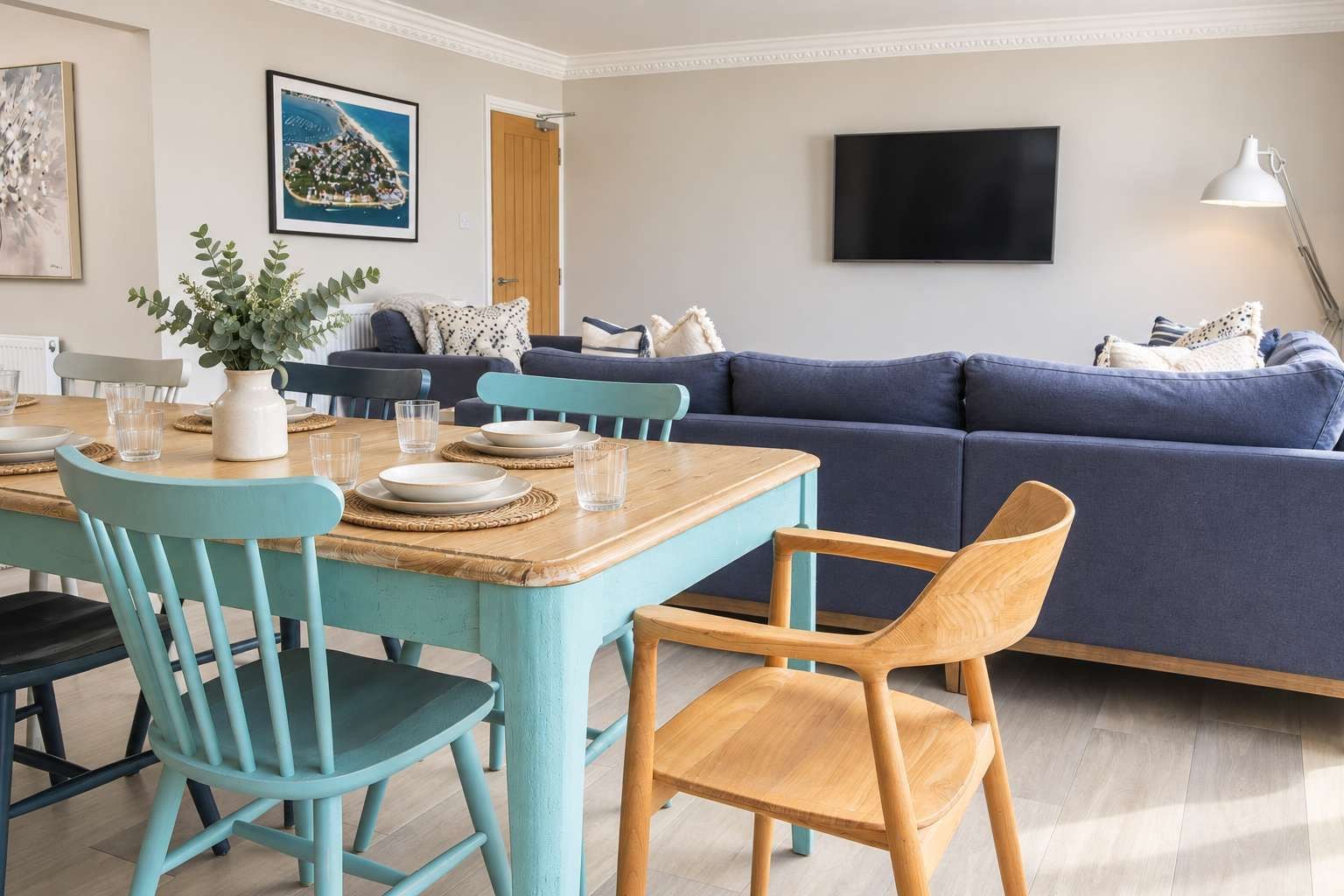 A modern living room with a wooden dining table, teal chairs, a blue sofa, and a flat-screen TV.