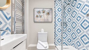 Et badeværelse med et hvidt toilet, en bruseniche med et geometrisk flisemønster og et billede i ramme af palmer.