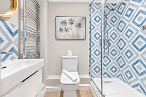 A bathroom with a white toilet, a shower with a geometric tile pattern, and a framed painting of palm trees. - High Tides Elegant 5-Bed Villa & Hot Tub Sandbanks (Sandbanks)