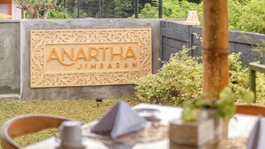 Μια πινακίδα με την επιγραφή «ANARTHA JIMBARAN», τοποθετημένη σε ένα διακοσμητικό ξύλινο πλαίσιο, περιτριγυρισμένη από πράσινο και έναν τοίχο από μπετόν.