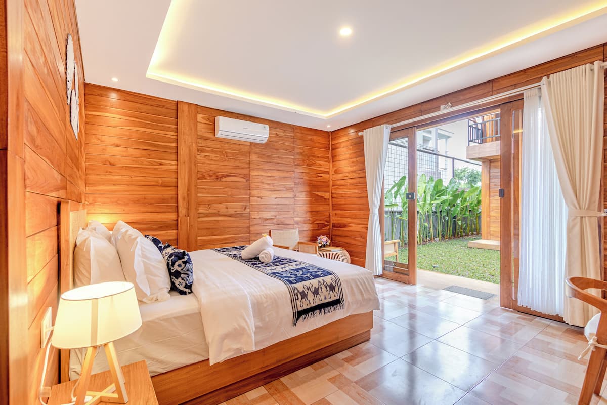 A wooden-paneled bedroom with a bed, a bedside lamp, and a view of a garden through a sliding glass door.