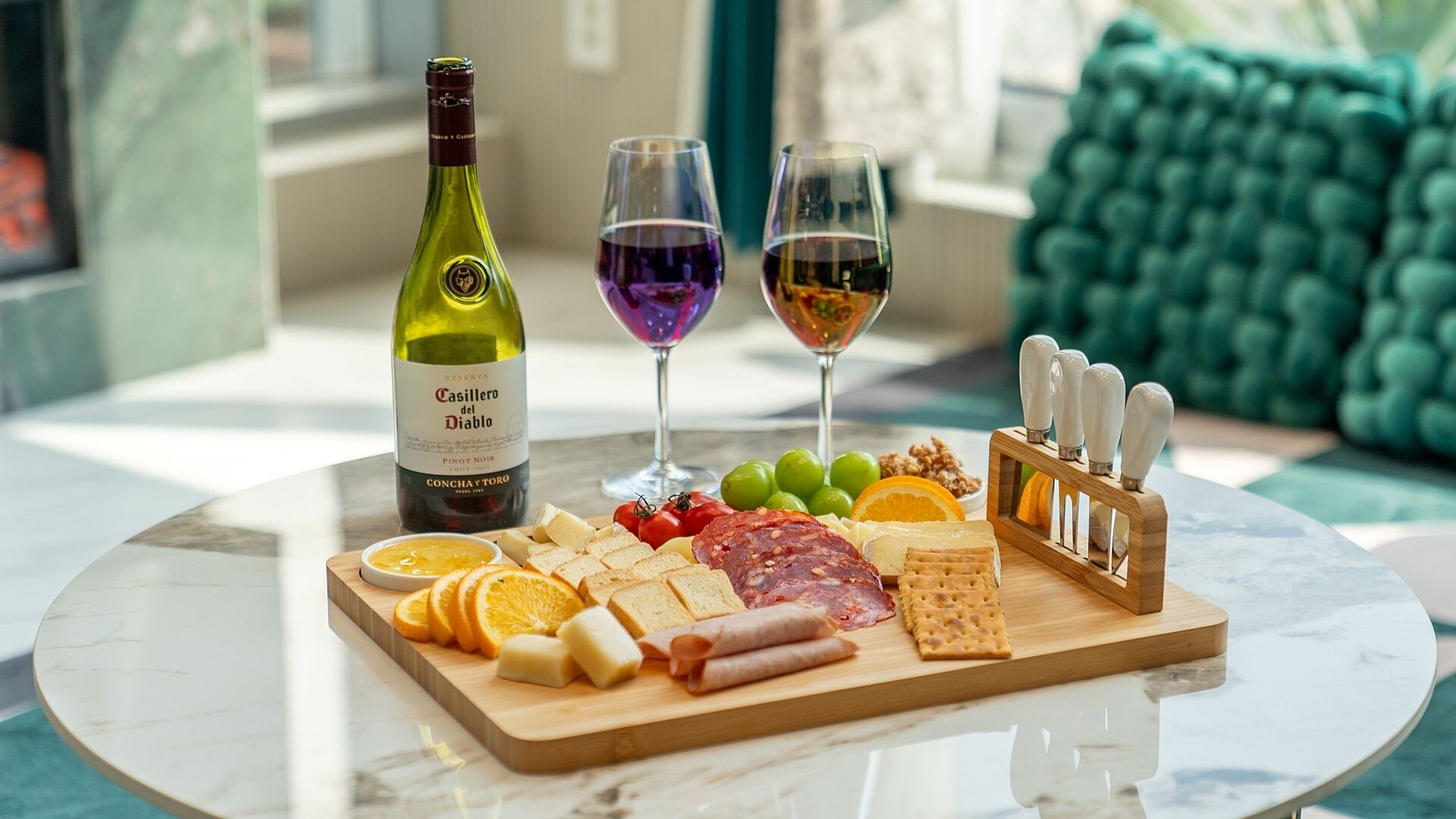 Een tafel met een fles Casillero del Diablo wijn, twee wijnglazen, een charcuterieplank met kaas, vleeswaren en fruit, en een messenblok.
