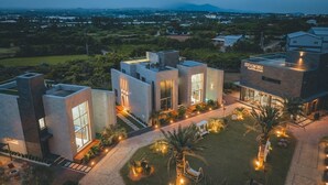 A modern hotel with a flat roof, large windows, and a well-lit outdoor area with palm trees and seating. - EUDAIMONIA (Jeju City)
