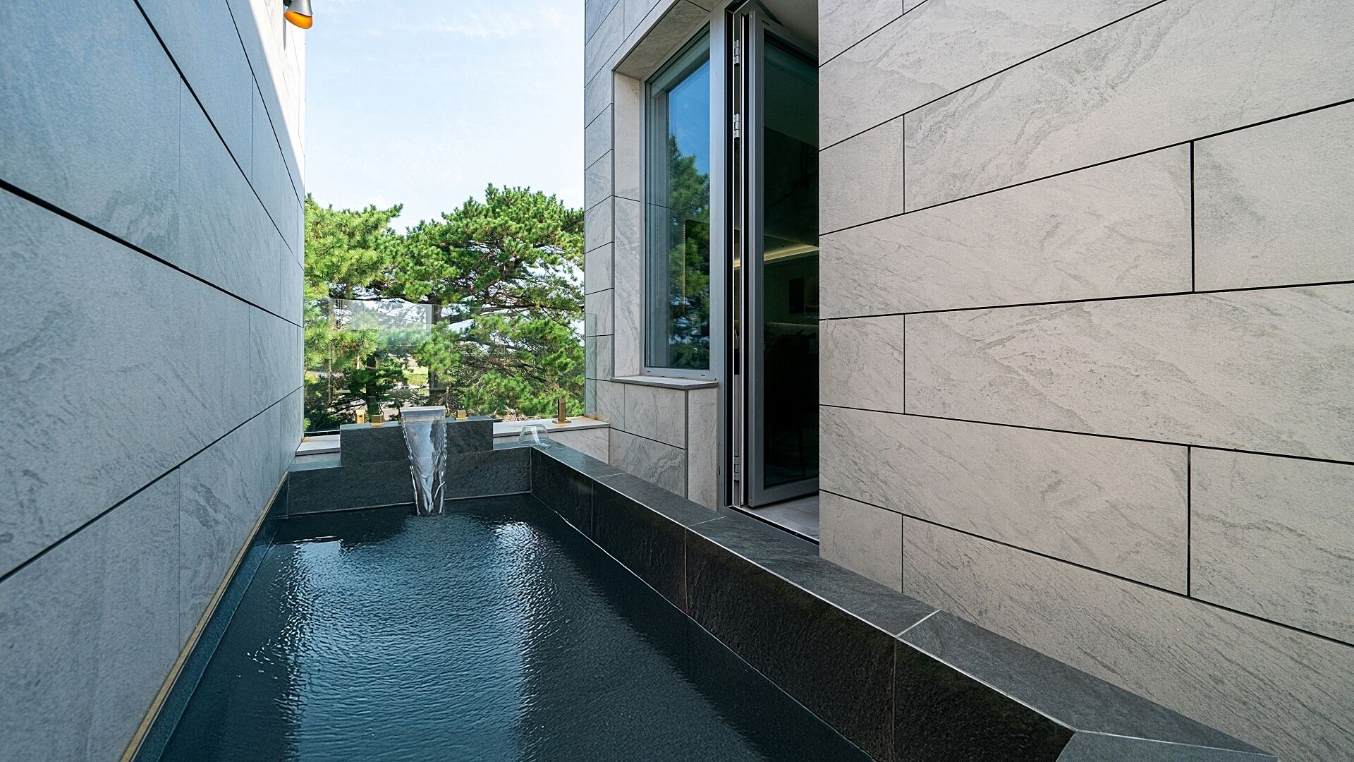A narrow outdoor water feature with a modern design, flanked by large stone tiles and a glass door.