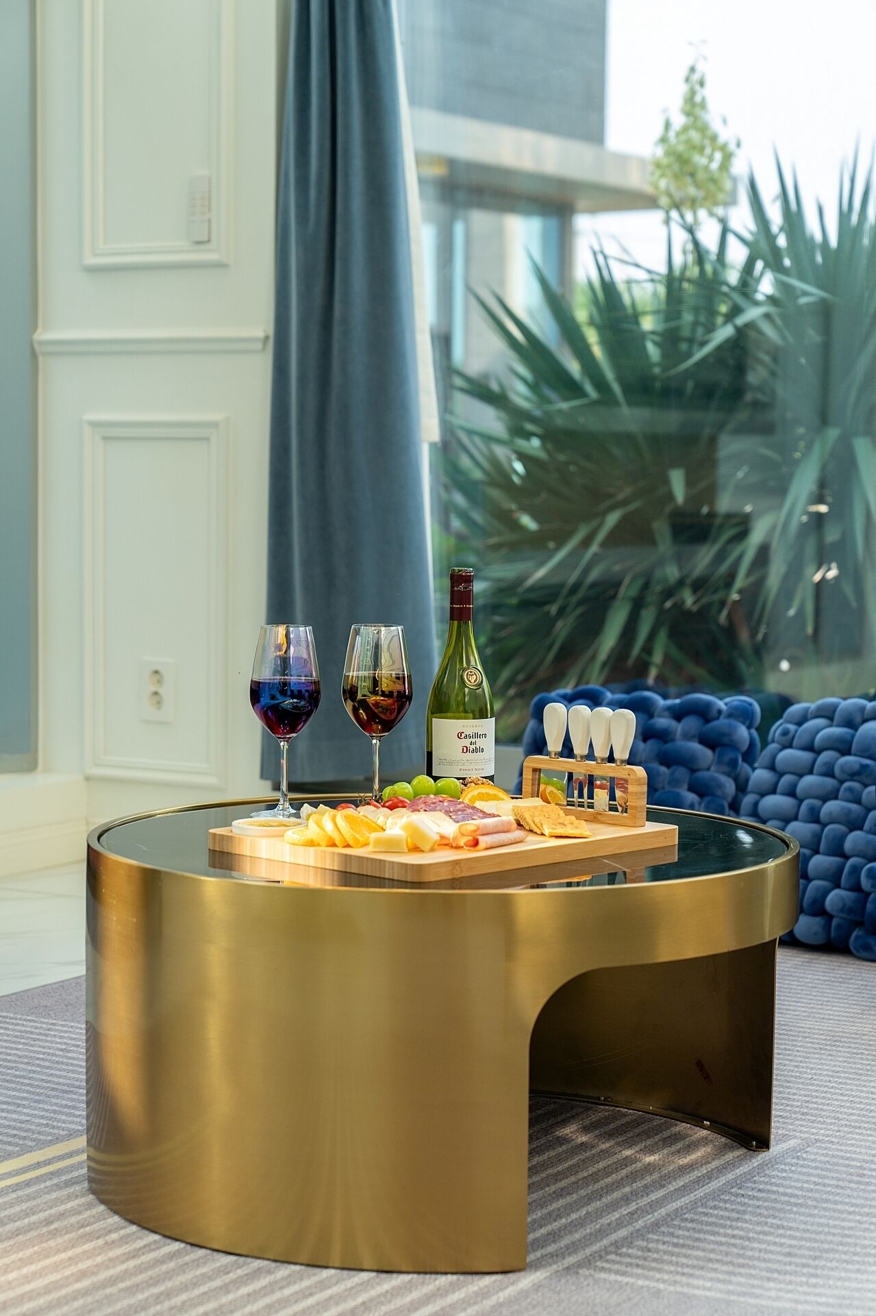 A round, gold-colored table with a cheese board, two glasses of red wine, and a bottle of Casillero del Diablo wine.