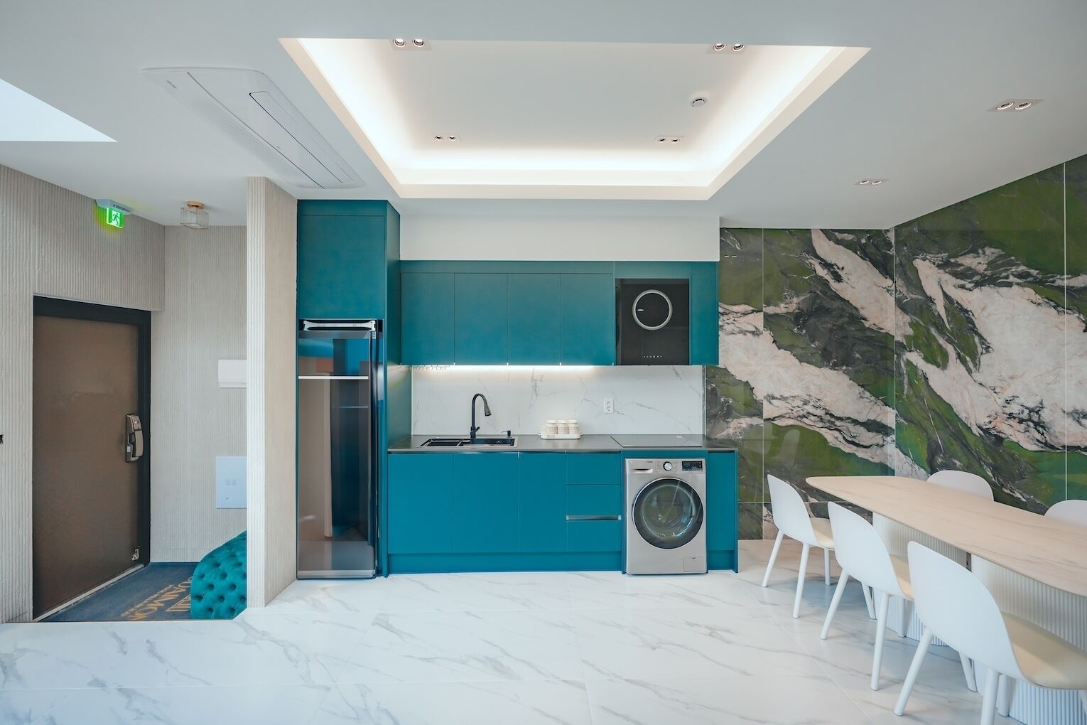 A modern kitchen with teal cabinetry, a built-in washing machine, and a marble backsplash.
