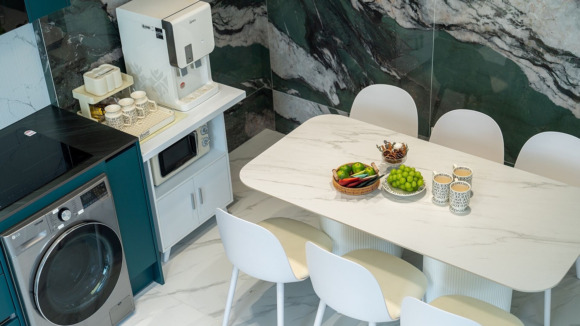 A modern kitchen with a built-in coffee maker, a washing machine, and a dining area with a marble table and white chairs.