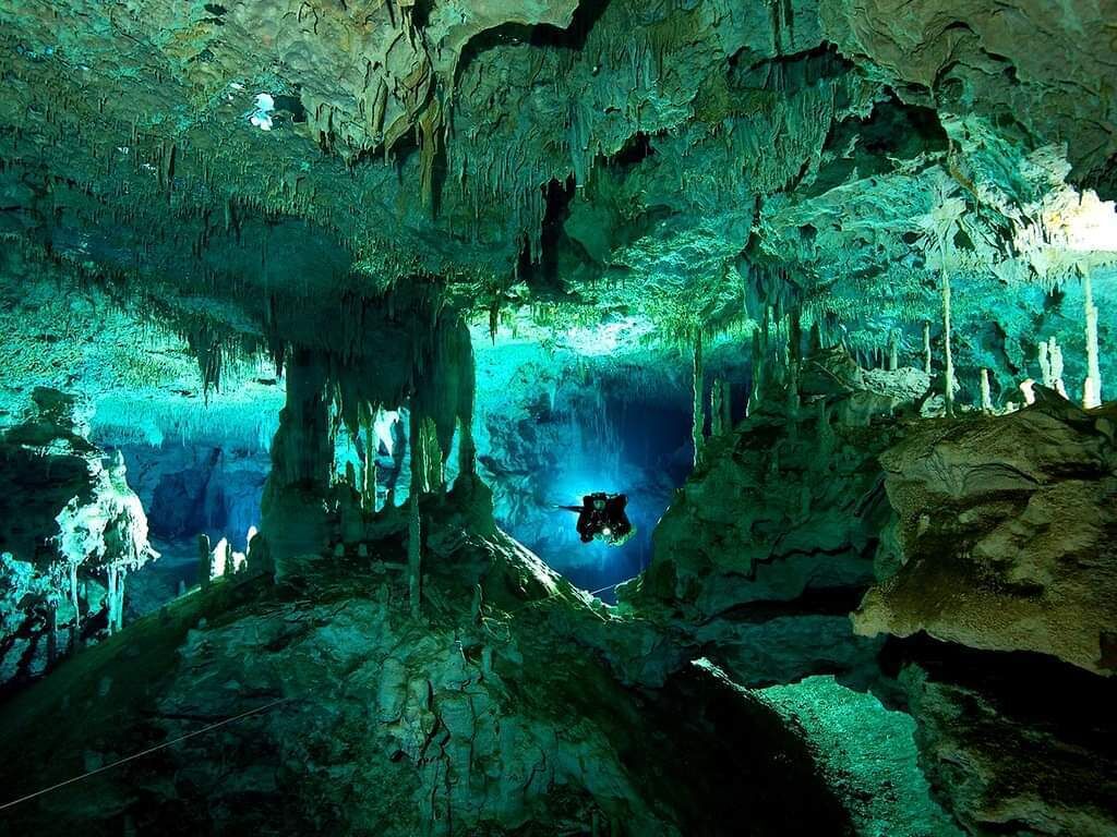 A cave with stalactites and stalagmites, a diver in the water, and a large rock formation.