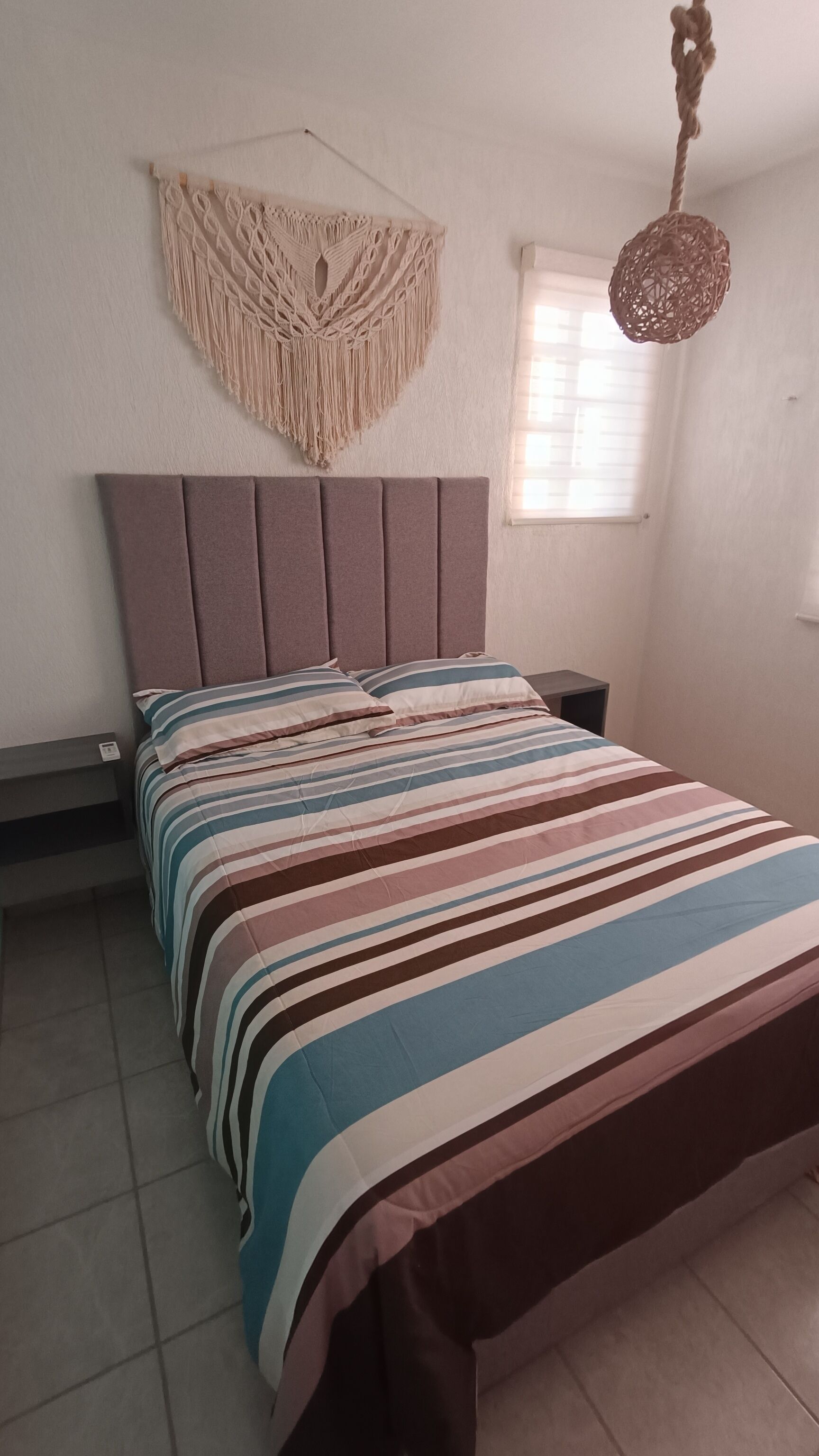 A bedroom with a bed, a macrame wall hanging, a woven light fixture, and a window with blinds.