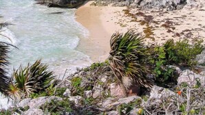 Vista costeira com uma grande estrutura de pedra, praia e terreno rochoso.