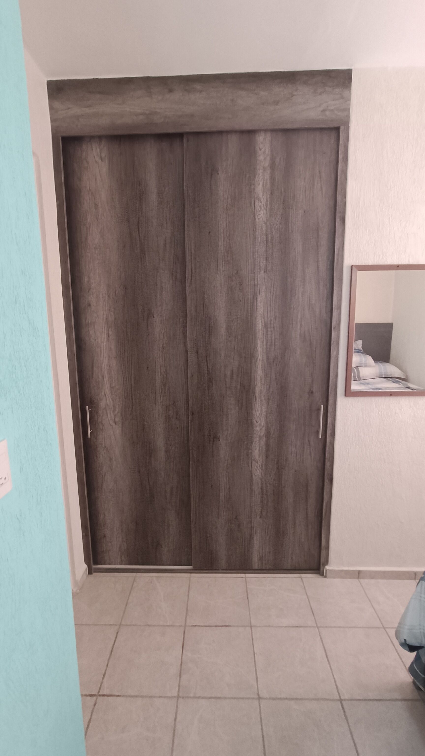 A room with a large wooden wardrobe, a tiled floor, and a small mirror on the wall.