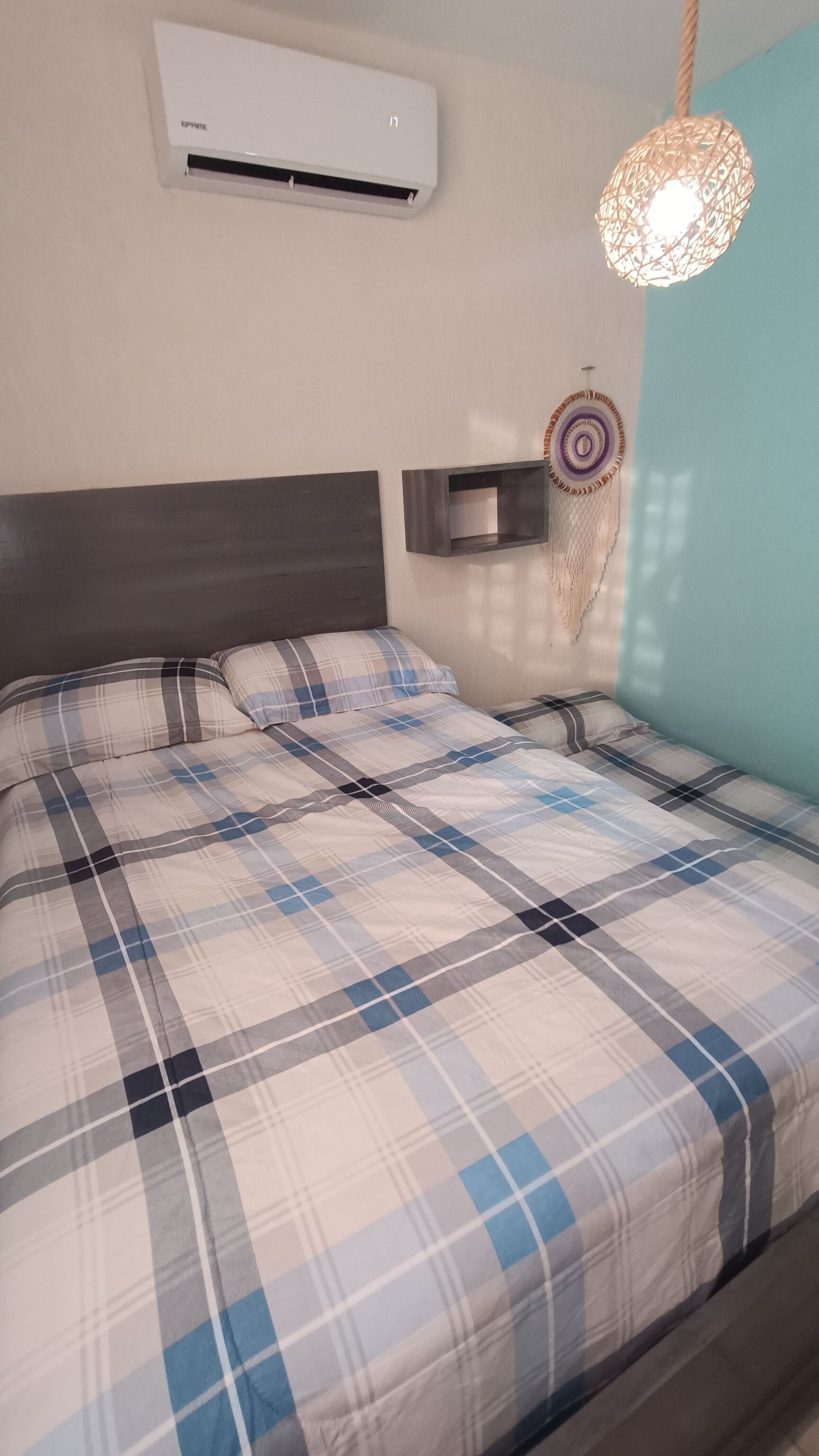 Ein Bett mit karierter Bettdecke, eine Wandklimaanlage, ein Wandregal und eine Hängeleuchte.