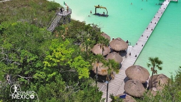 Um resort costeiro com um cais que se estende por águas turquesa, cercado por vegetação exuberante e cabanas com telhado de palha.