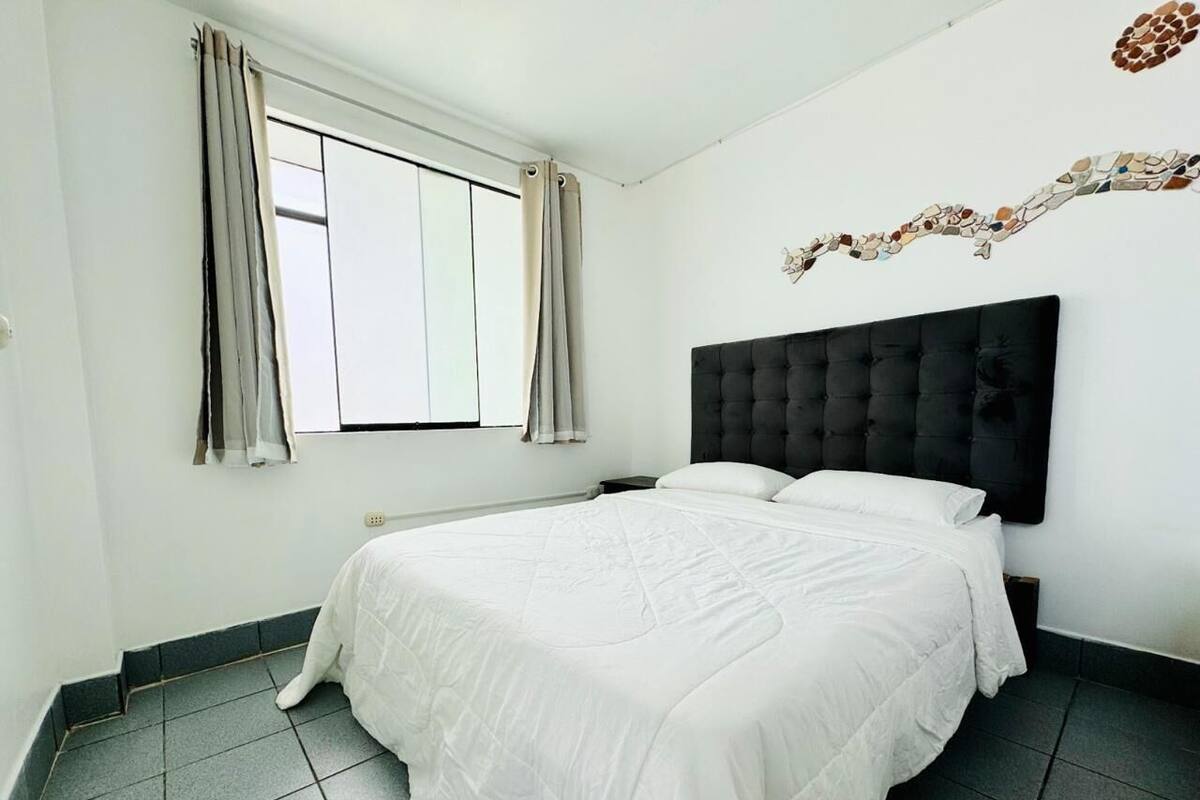 A bedroom with a large bed, a headboard, a window with curtains, and a wall decoration.