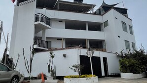 A multi-story hotel with a prominent water tank on the roof, multiple balconies, and a parking area with a speed limit sign. - Hotel La planicie -Pico Alto (Punta Hermosa)