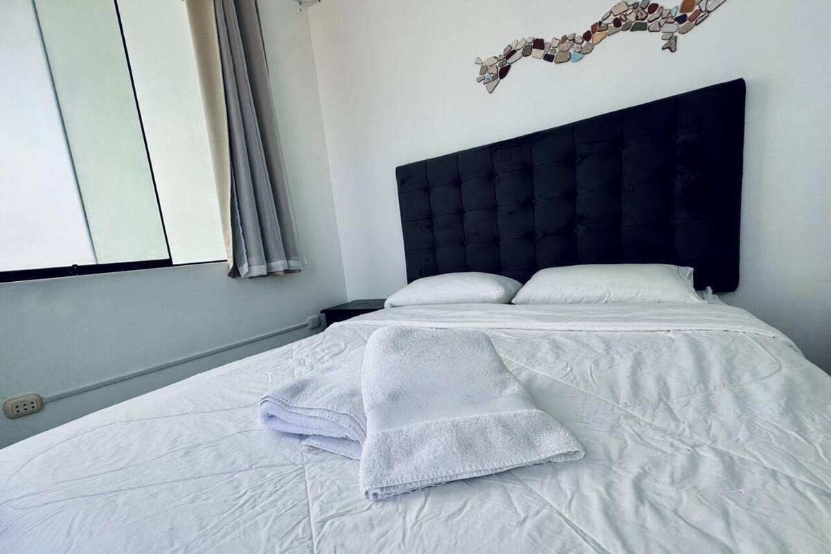 A neatly made bed with white linens and a folded towel, a dark headboard, and a window with curtains.