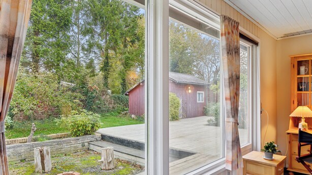 A room with a large window, a wooden desk, a lamp, and a view of a red barn and greenery outside. - Cozy summer house by Stevns Klint and Rødvig (Rødvig Stevns)