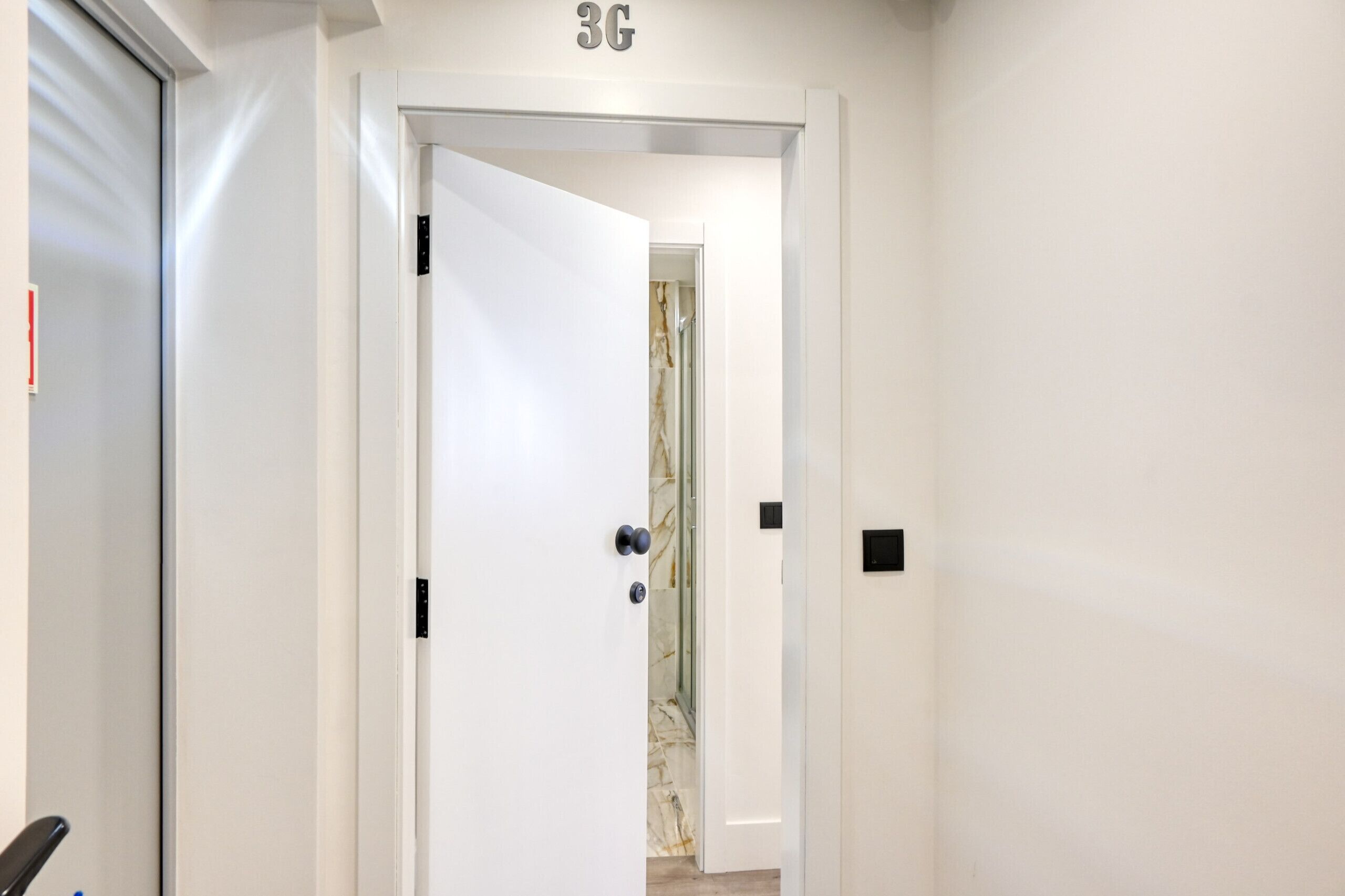 A hallway with a white door marked "3G" leading to a room with a marble wall and a shower.