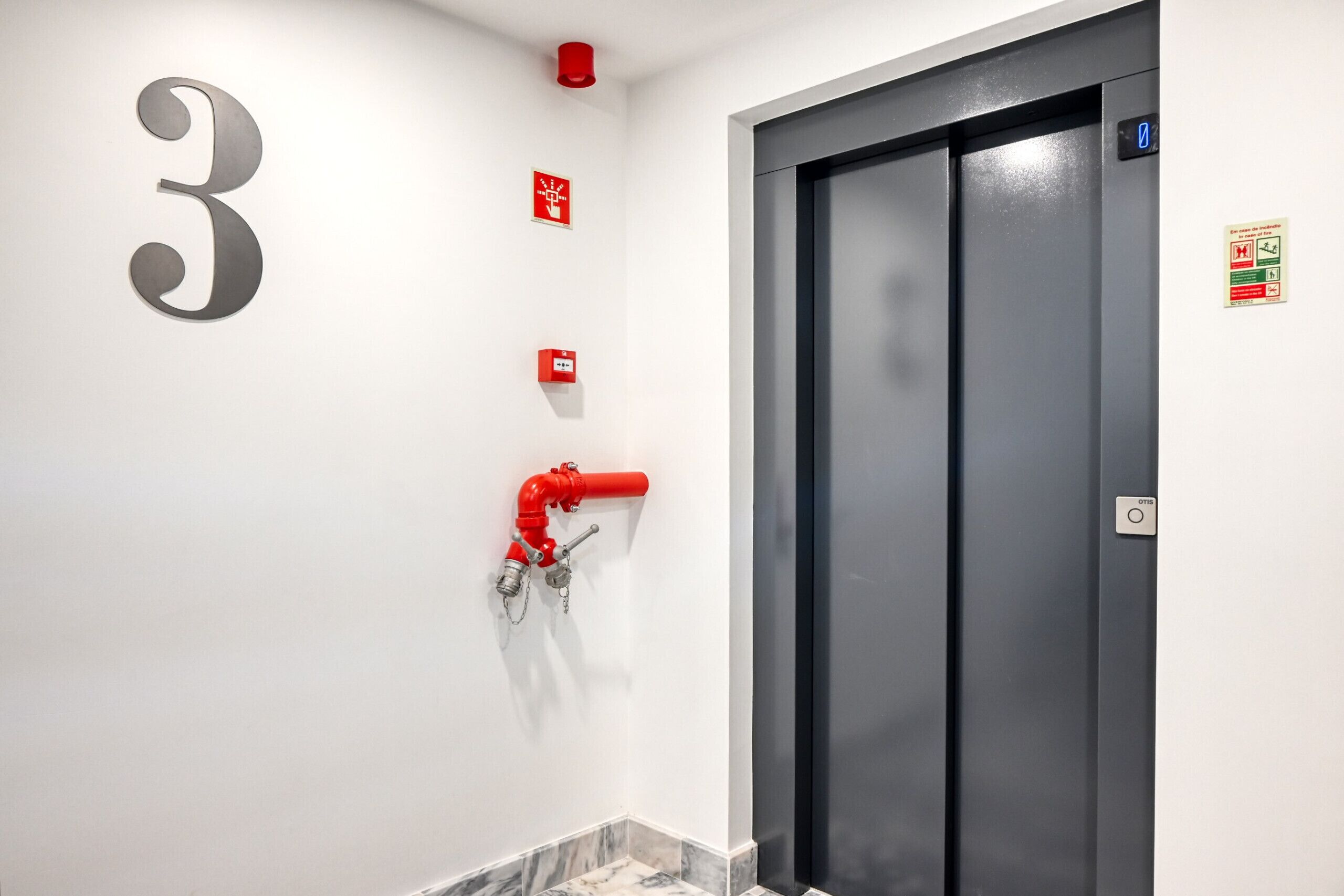 Un ascenseur moderne avec une porte sombre, un panneau indiquant le numéro « 3 » sur le mur et un tuyau d’incendie rouge sur le sol.