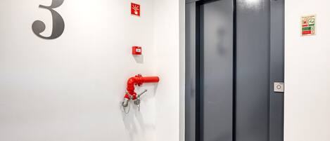 Un ascenseur moderne avec une porte sombre, un panneau indiquant le numéro « 3 » sur le mur et un tuyau d’incendie rouge sur le sol.