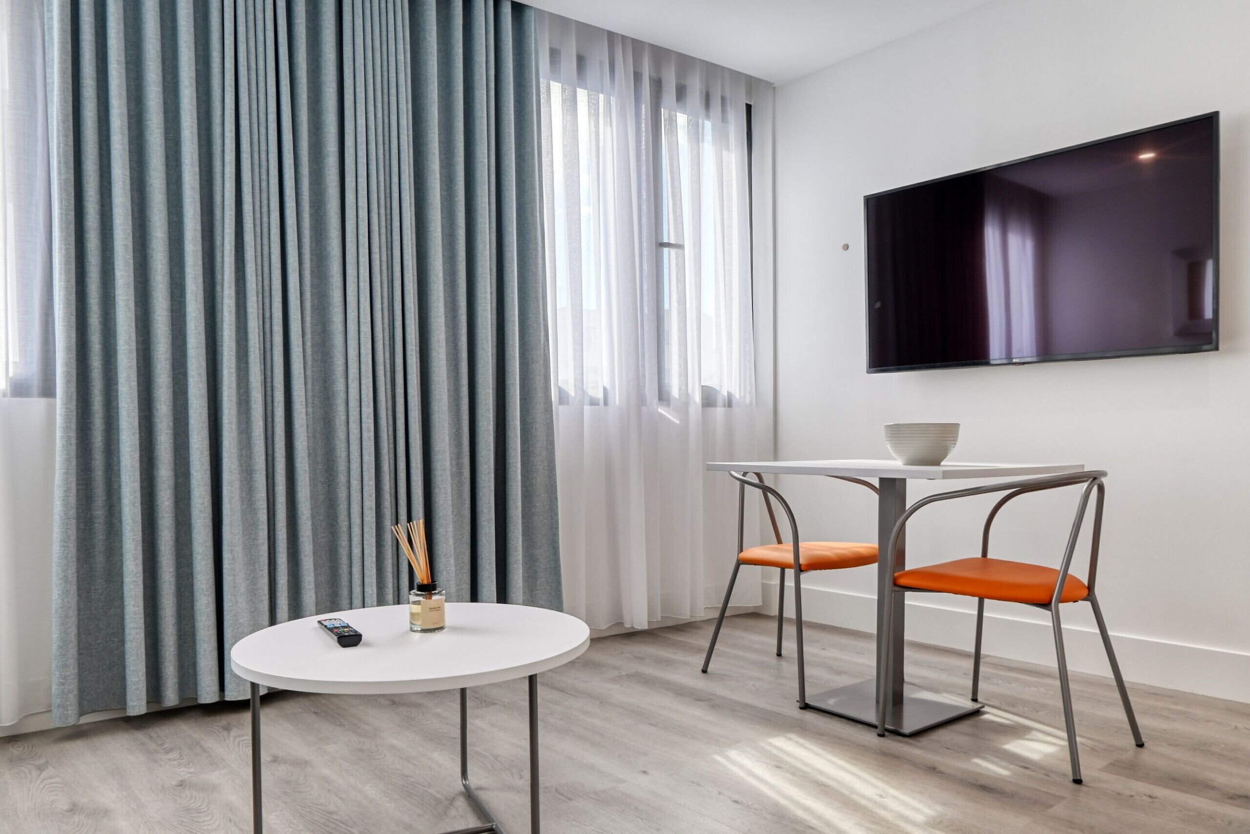Ein modernes Zimmer mit rundem Tisch, zwei Stühlen, einem Fernseher an der Wand und einem großen Fenster mit Vorhängen.