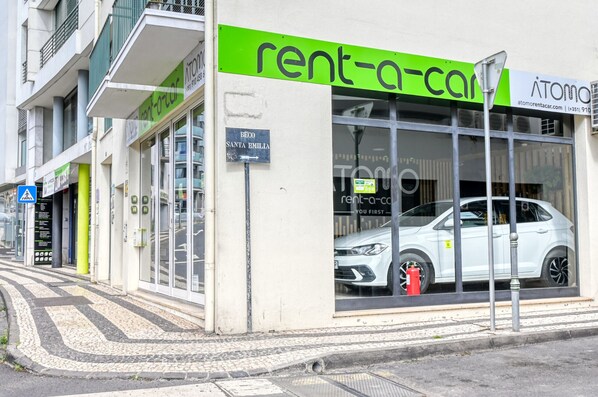 Un servicio de alquiler de coches con un amplio escaparate que muestra un coche blanco, un cartel que dice 'rent-a-car' y un cartel con el nombre 'Bego Santa Emilia'.