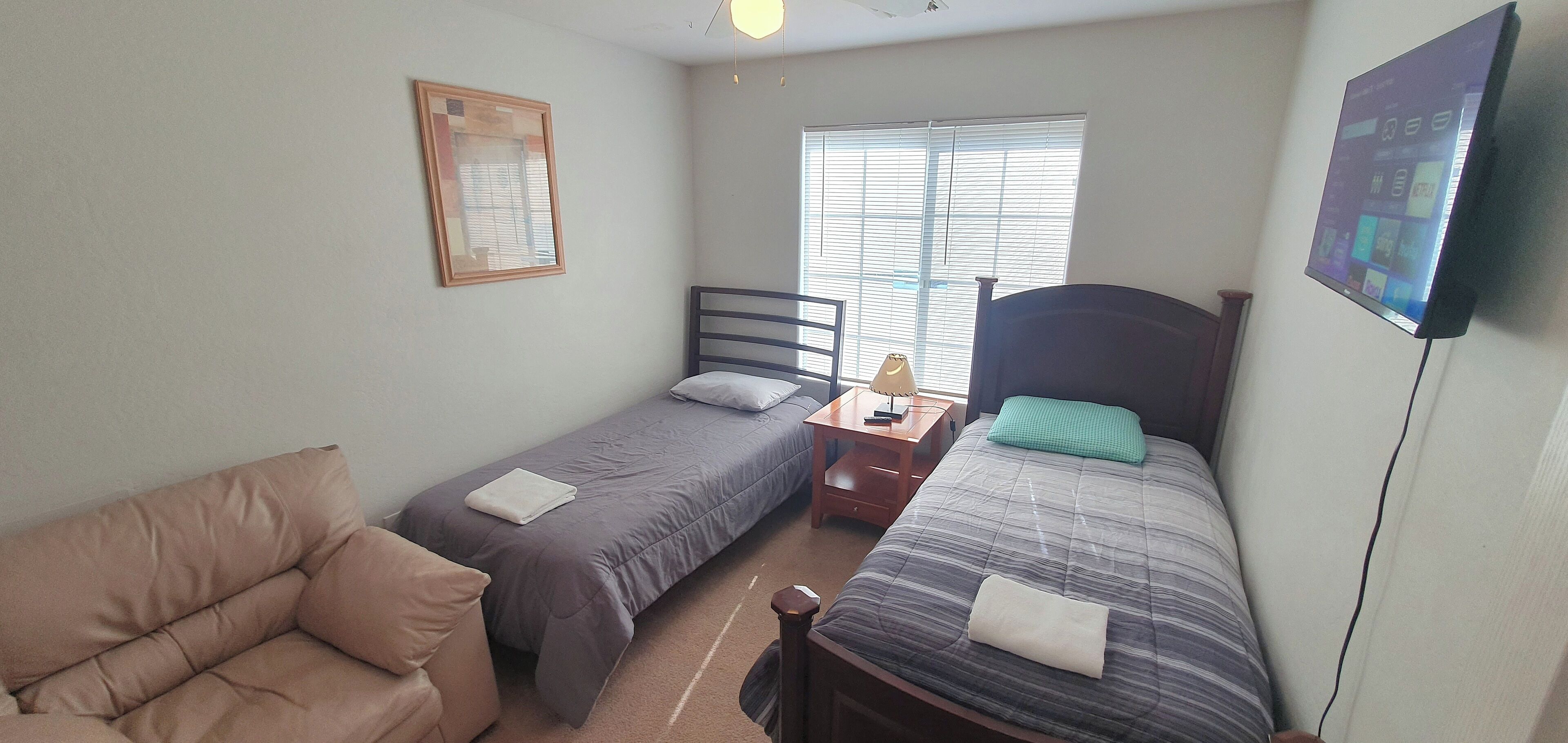 A small, well-lit room with two single beds, a sofa, a nightstand with a lamp, and a TV mounted on the wall.
