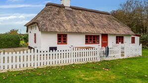 A traditional thatched-roof house with white walls and red doors, surrounded by a white picket fence. - Awesome home in Tim with WiFi (Tim)