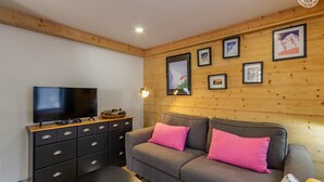 A cozy living room with a flat-screen TV, a wooden cabinet, and a grey sofa with pink cushions.