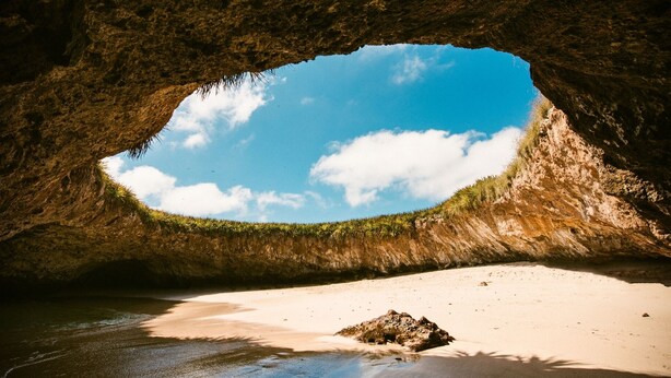A natural cave opening with a sandy beach and a clear blue sky visible through the arch.