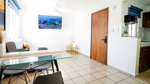 A dining area with a glass table, chairs, a wooden door, and a kitchen visible through an open doorway.