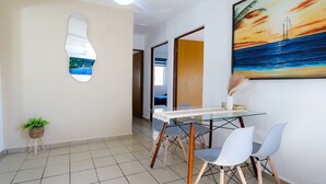 A dining area with a glass table, white chairs, a potted plant, and a painting of a sunset over the ocean.
