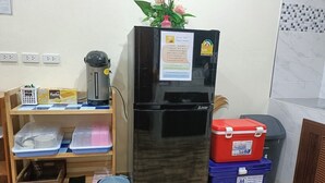 A kitchen with a black refrigerator, a wooden shelf with kitchenware, a trash bin, and a blue storage bin. - Saendee Pool Villa (Klaeng)