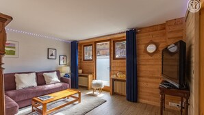 A cozy living room with a wooden floor, a purple sofa, a wooden coffee table, a television, and a large window with curtains. - La Duchere - 1st Floor (LA GIETTAZ)