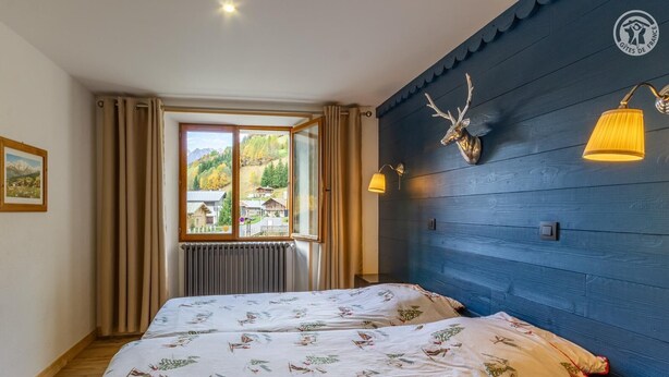 A bedroom with a bed, a window with curtains, a radiator, a wall-mounted deer head decoration, and two wall-mounted lamps. - La Duchere - 1st Floor (LA GIETTAZ)