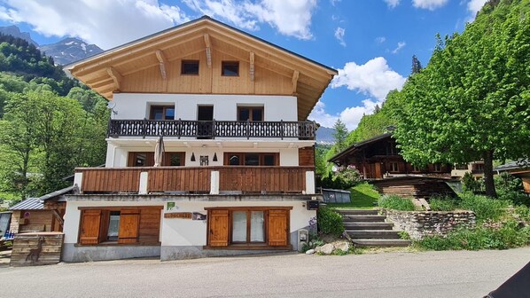 Un chalet à deux étages doté de balcons en bois, d’un escalier en pierre et surplombant un paysage montagneux.