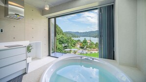 A modern bathroom with a bathtub, a toilet, and a sink. It features a large window with a view of a lake and mountains.