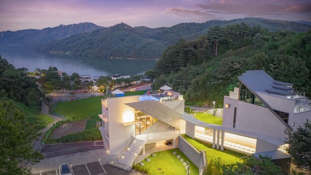 A modern building with a glass facade, surrounded by lush greenery and a lake.