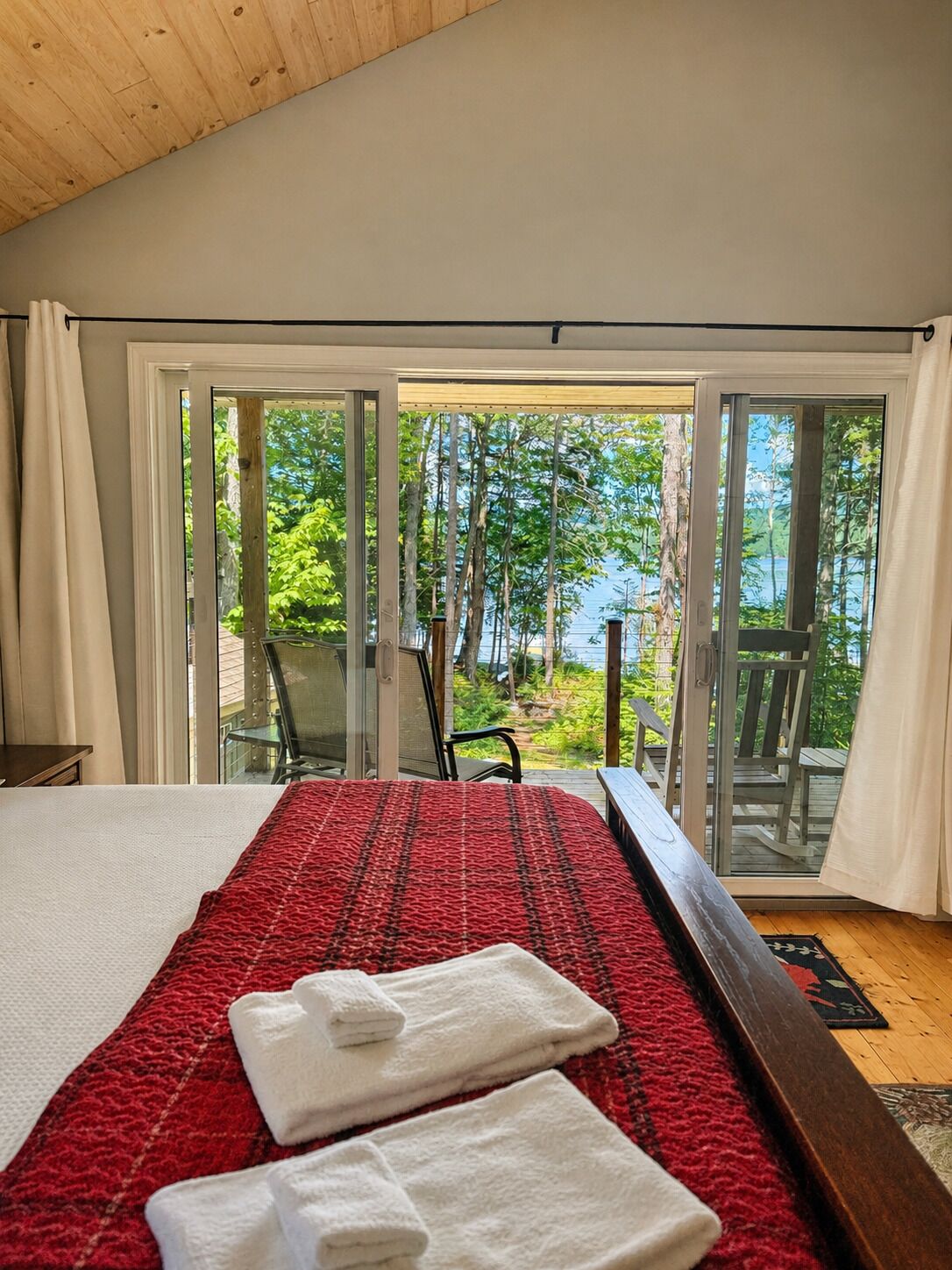 A bedroom with a large bed, a view of a forest through glass doors, and a wooden ceiling.