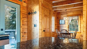 L’intérieur d’un chalet en bois est doté d’un plan de travail en granit, de murs en bois, d’une table à manger avec des chaises et d’une fenêtre offrant une vue sur des arbres couverts de neige.