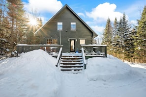A log cabin with a wooden staircase leading to a front door, surrounded by snow and trees.
