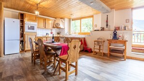 Une cuisine spacieuse avec des meubles en bois, une table à manger avec des chaises, une nappe rouge et une vue sur l’extérieur grâce à de grandes fenêtres.