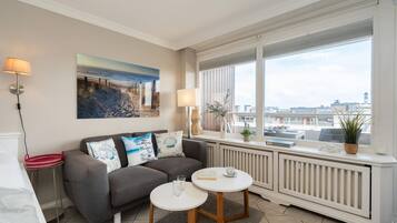 Ein modernes Wohnzimmer mit einem grauen Sofa, zwei kleinen runden Tischen, einem großen Fenster mit Blick auf die Stadt und einem Gemälde an der Wand.