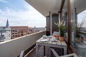 Un balcón con mesa lista para dos, una planta en maceta y vista a edificios al fondo.
