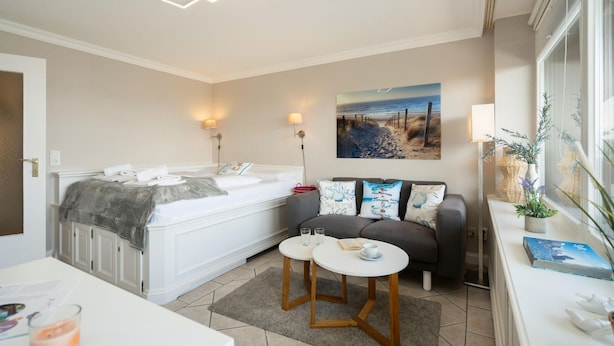 A modern bedroom with a large bed, bedside lamps, a painting of a beach, a small table with a book and glasses, and a window with plants. - Küstenmuschel - Modern 1-room apartment, close to the beach, balcony, WLAN. Pets (Westerland)