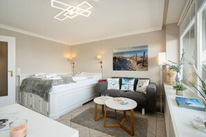 Een moderne slaapkamer met een groot bed, nachtlampjes, een schilderij van een strand, een klein tafeltje met een boek en brillen, en een raam met planten.
