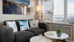 A modern living room with a grey sofa, three decorative pillows, a round white table with a book and a potted plant, a floor lamp, and a large painting of a beach scene. - Küstenmuschel - Modern 1-room apartment, close to the beach, balcony, WLAN. Pets (Westerland)